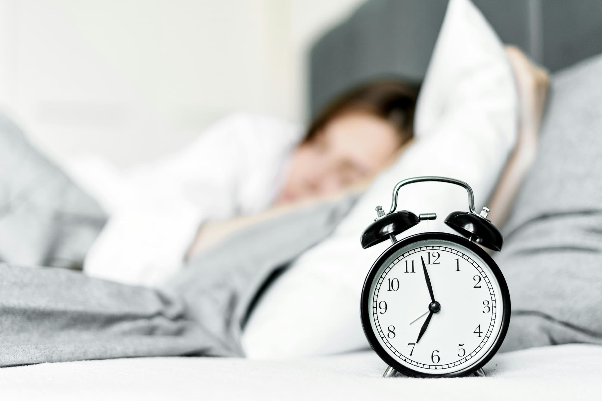 Classic alarm clock in focus with woman sleeping in the background, symbolising healthy sleep schedule and consistent bedtime routine.