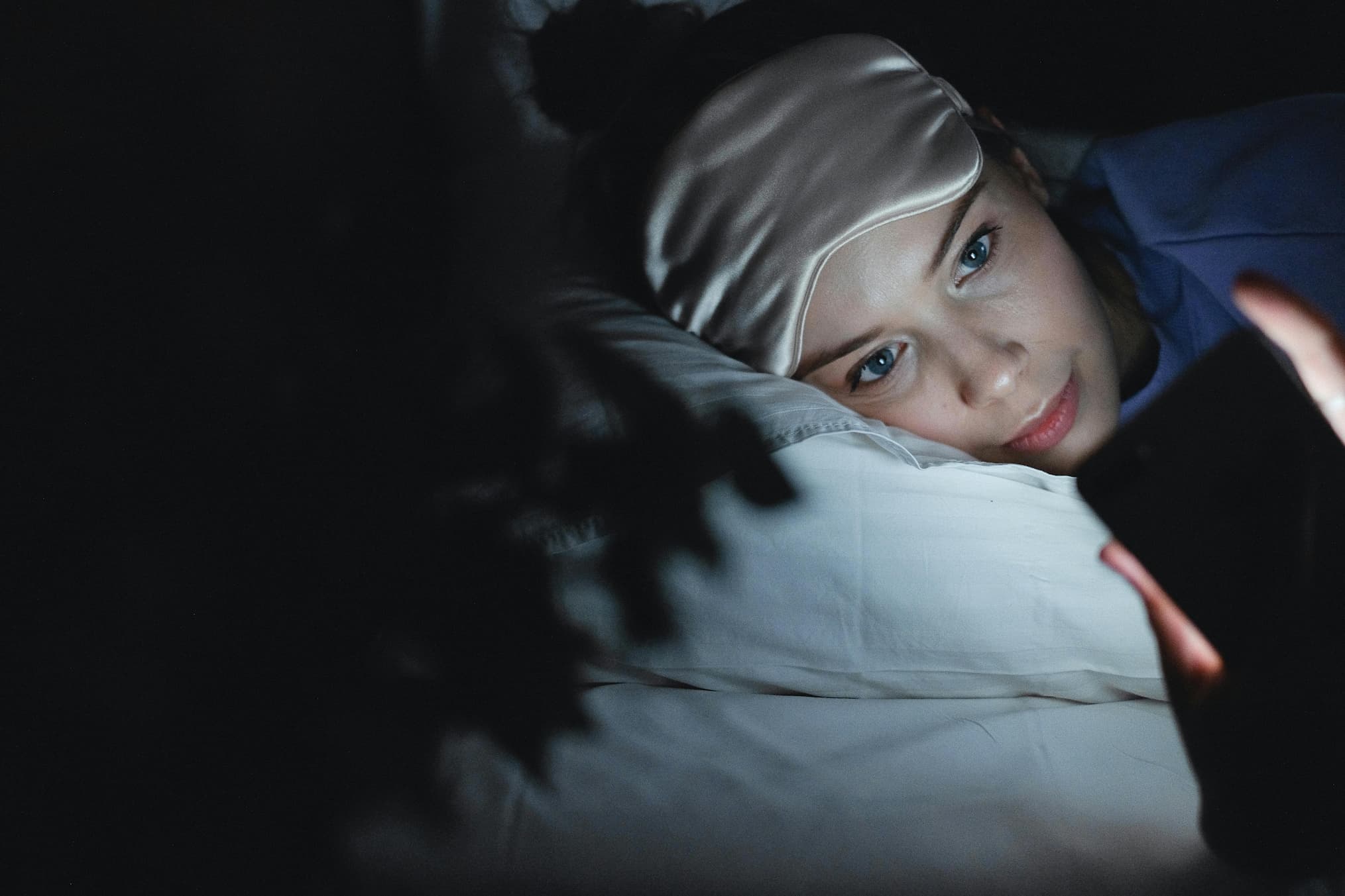 Woman lying in bed at night with a silk sleep mask pushed up, looking at her phone, highlighting the impact of screen time on sleep quality and melatonin levels.