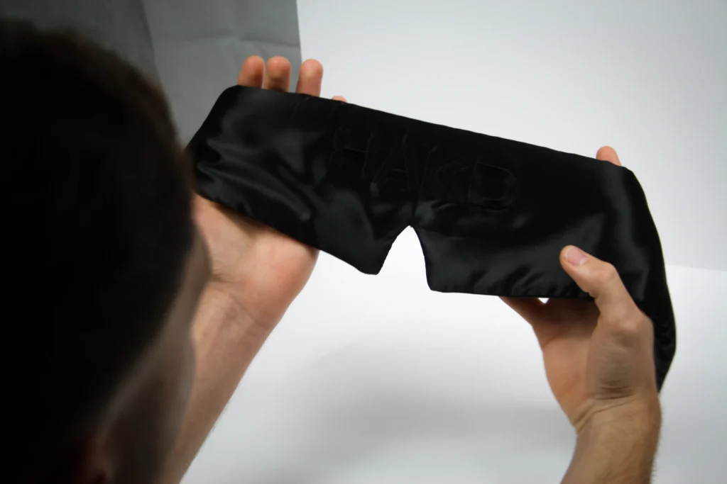 Man holding the HAKD Deep Sleep Mask, showcasing its sleek 100% blackout silk design for optimal comfort and light blocking.