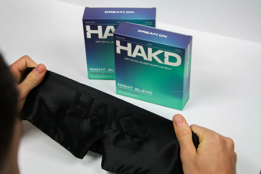 HAKD Night Blend sleep supplement boxes with a person holding a black HAKD silk sleep mask, highlighting a complete sleep optimisation setup.
